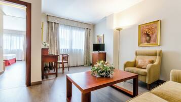 Habitación de hotel con piso de madera, un escritorio con silla, un sofá, una mesa de centro con flores, un televisor y un cuadro en la pared.
