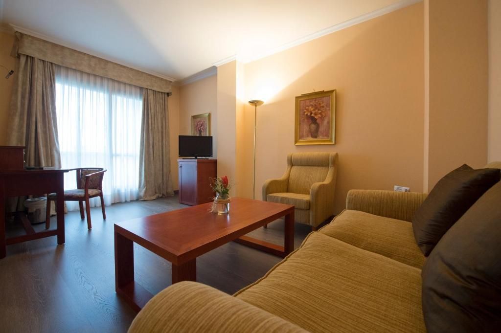 Habitación de hotel con sofá, una mesa de centro de madera, televisor, escritorio con silla y una ventana grande con cortinas.