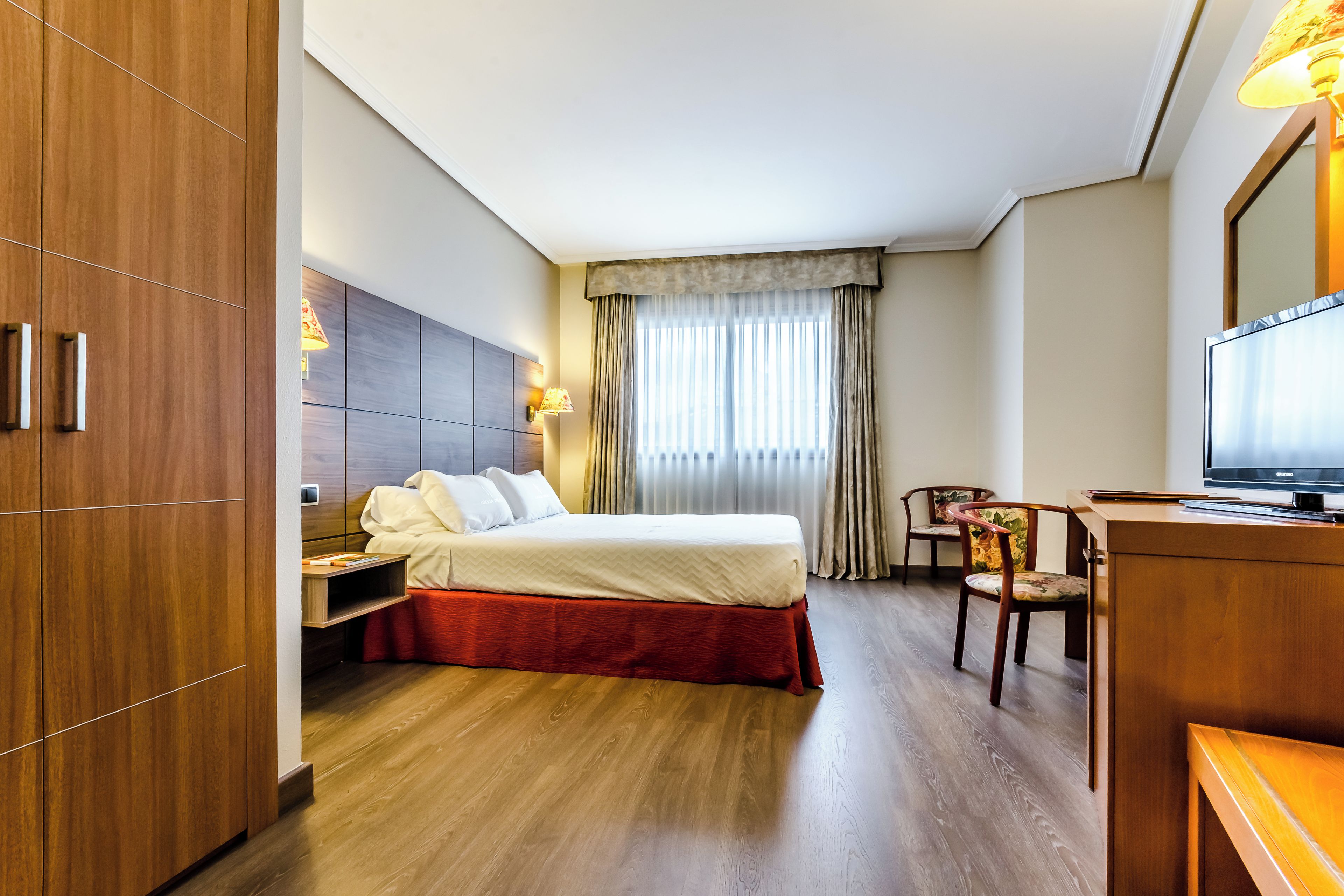 Quarto de hotel com uma cama, uma escrivaninha com televisão, uma cadeira e um guarda-roupa.