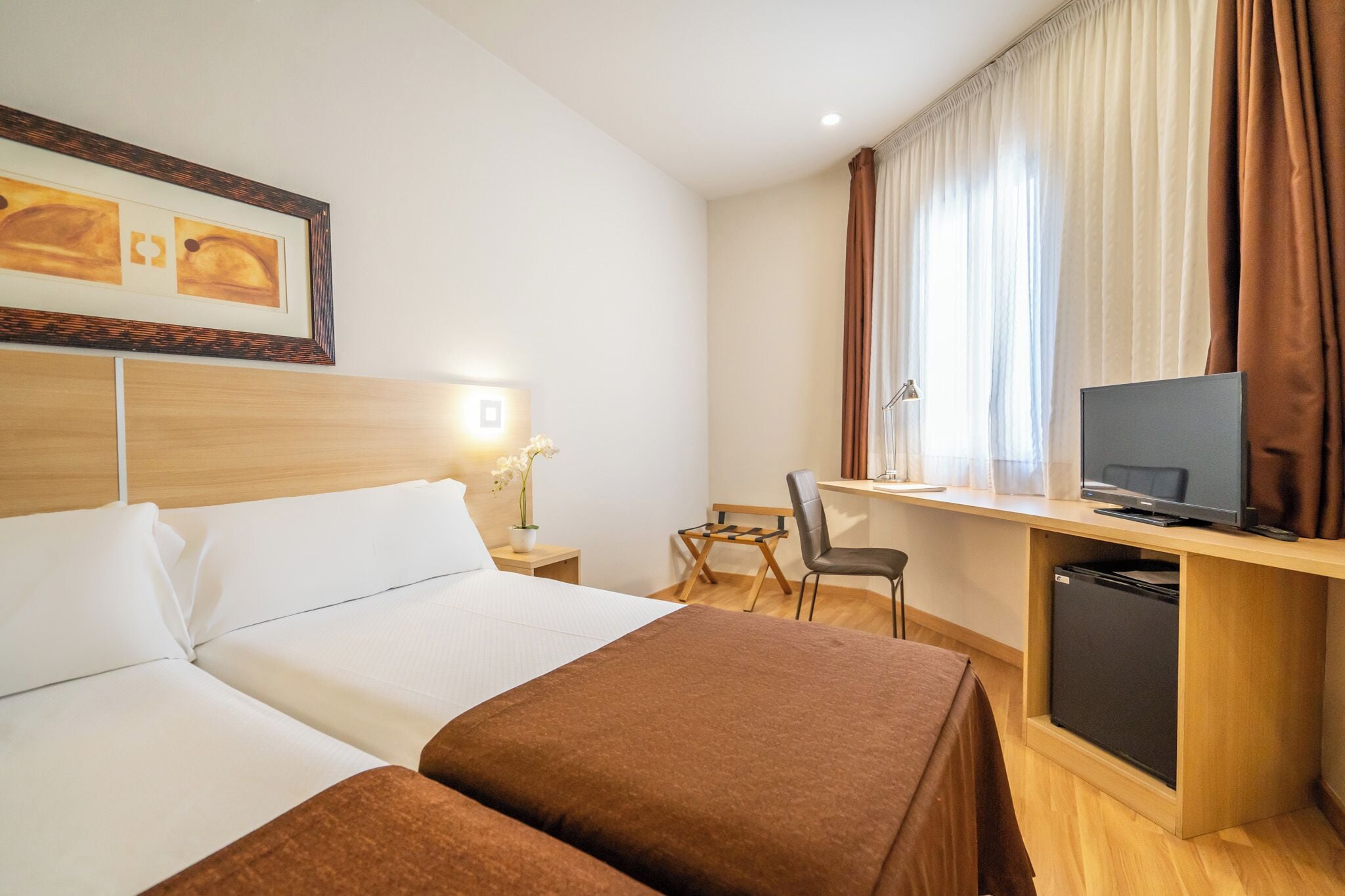 A hotel room with two beds, a desk with a television, a small table with a chair, and a window with curtains.