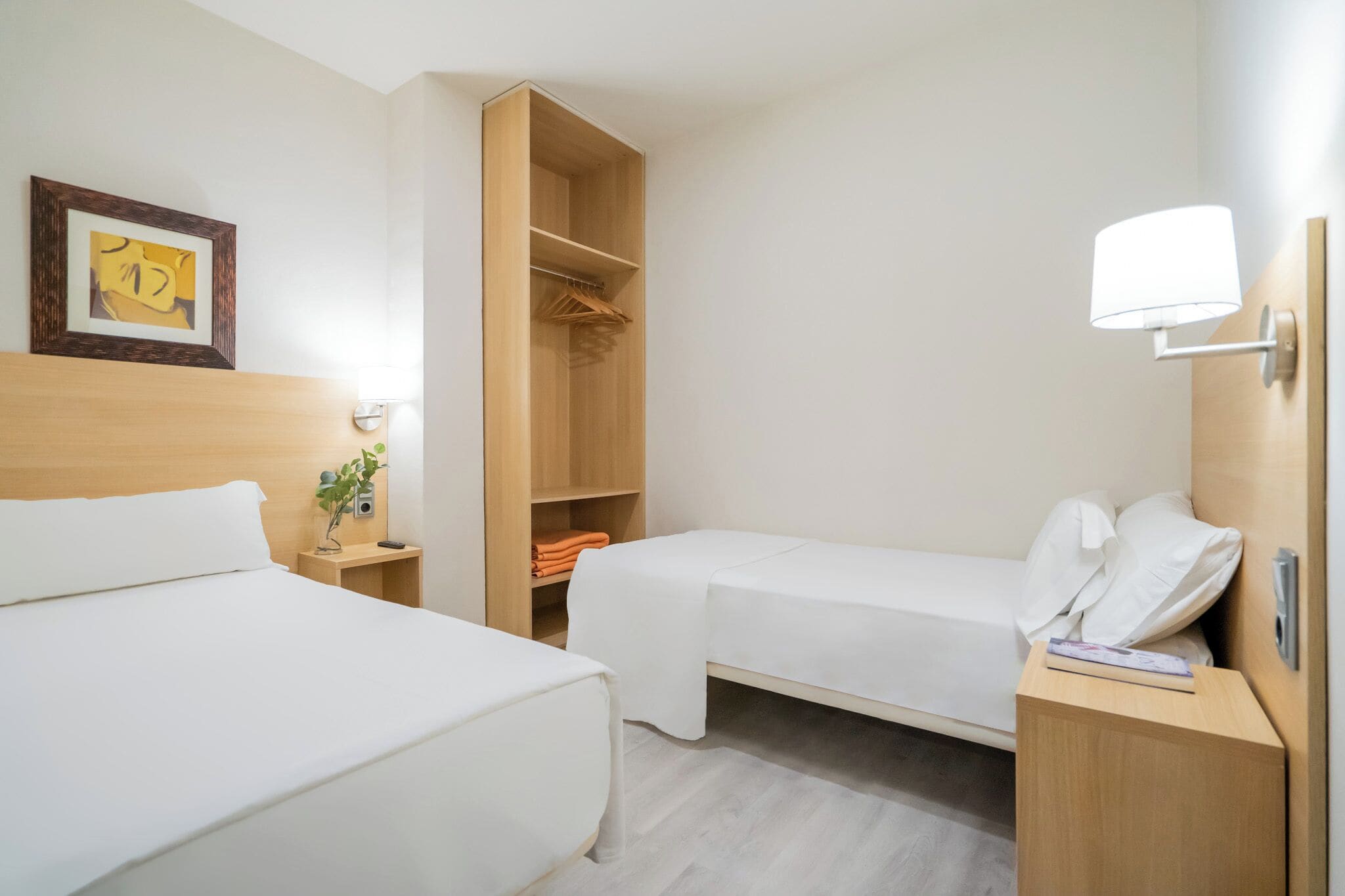 Une chambre d’hôtel avec deux lits simples, une armoire en bois, une table de chevet avec une lampe et un tableau encadré au mur.