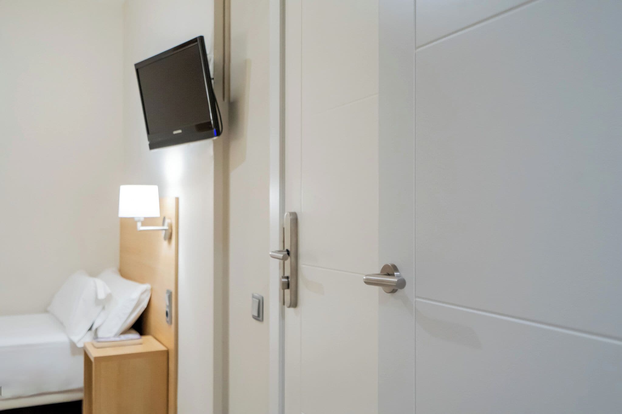 A hotel room with a bed, a bedside table, a TV mounted on the wall, and a door with a handle.