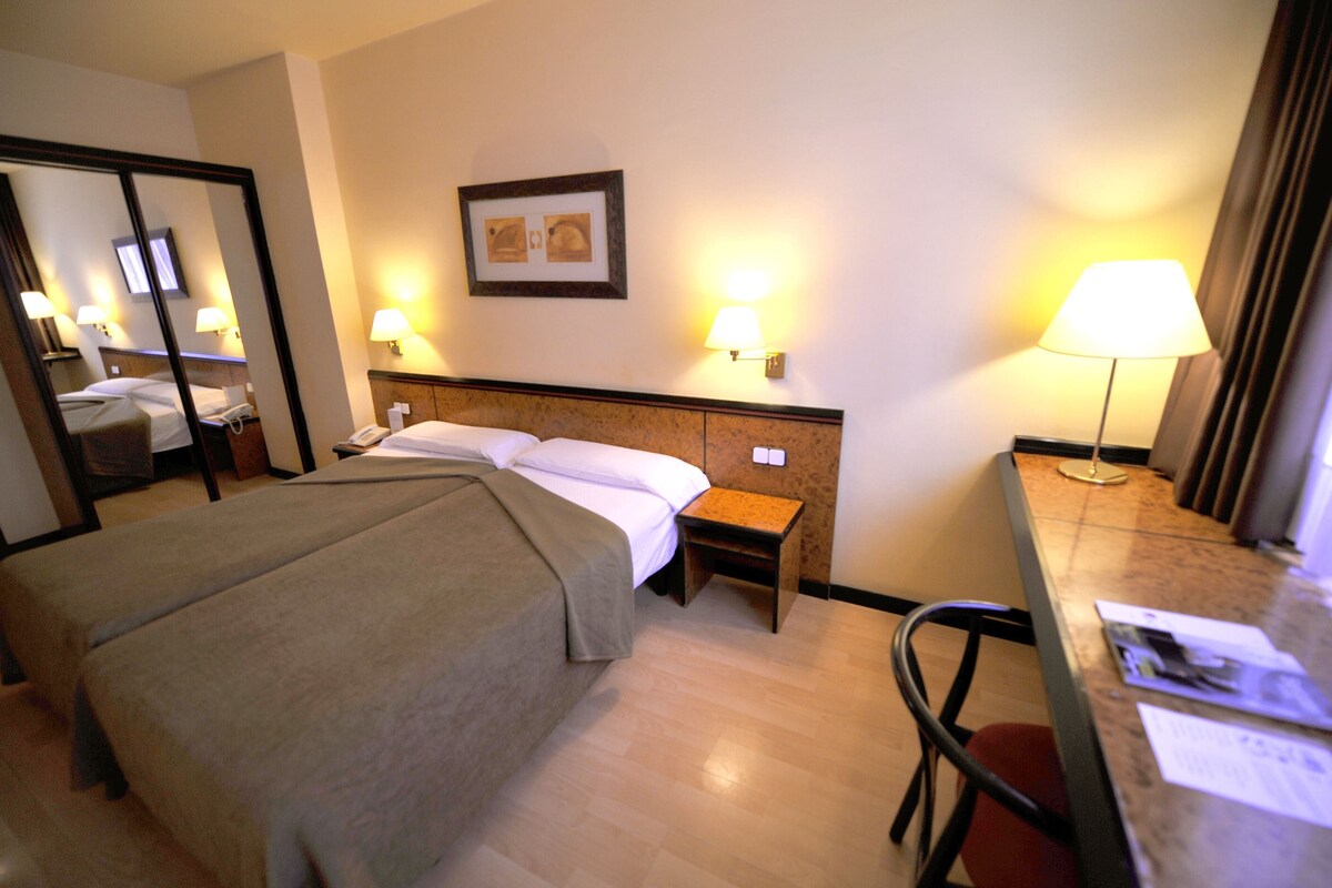 Una camera d'albergo con un letto, comodino, lampada, sedia e una scrivania illuminata da una lampada.