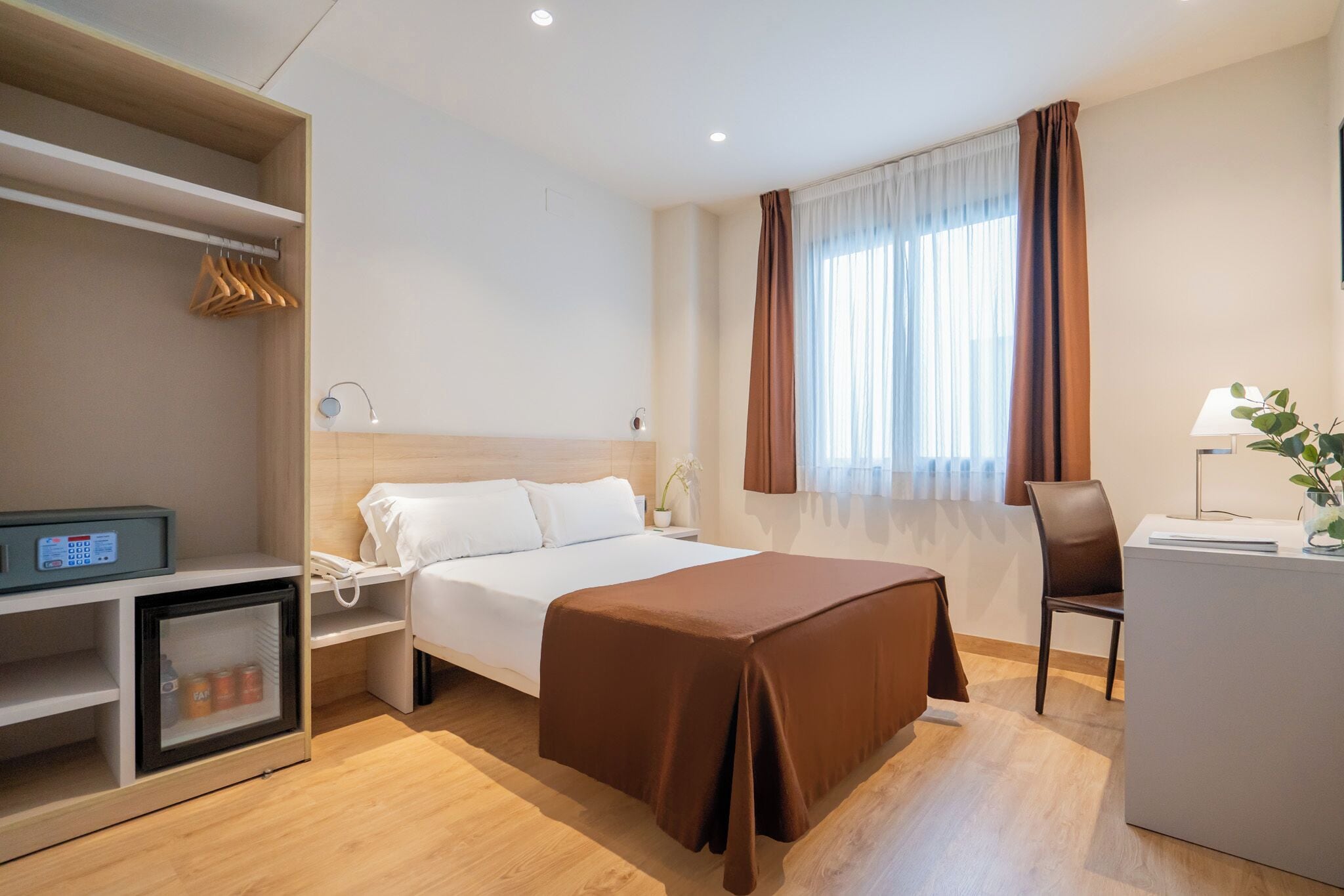 A hotel room with a bed, bedside table, desk, chair, and a minibar.