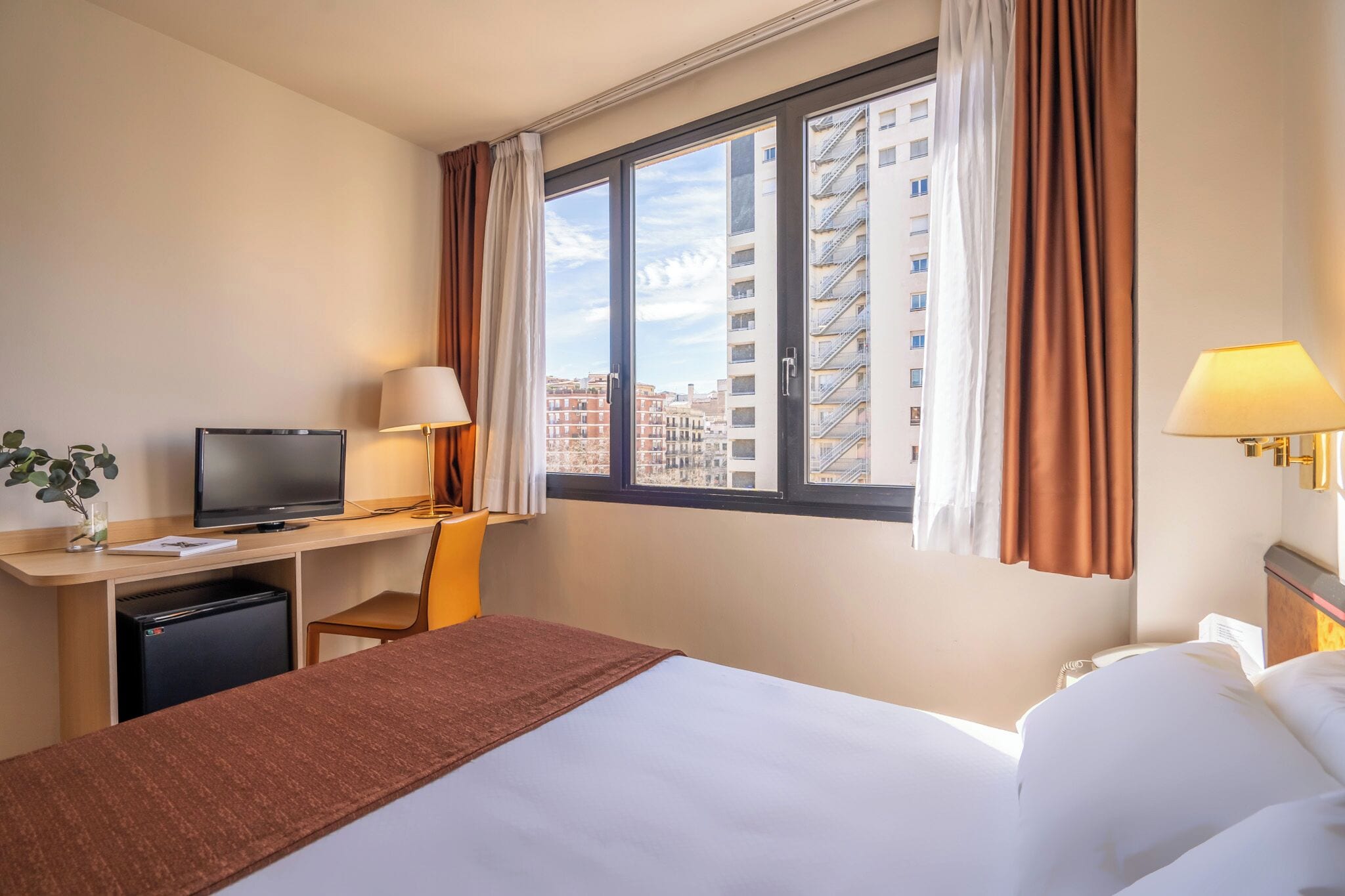 Habitación de hotel con cama, escritorio, televisión y ventana con cortinas.