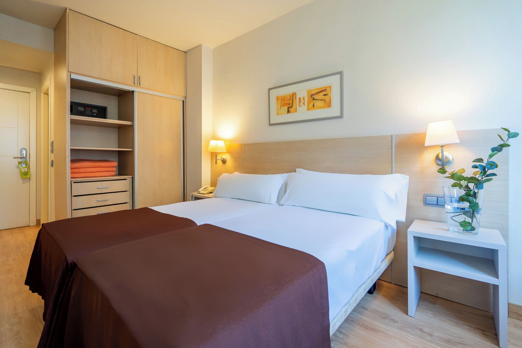 A hotel room with a large bed, a wardrobe, a bedside table with a lamp, and a wall-mounted artwork.
