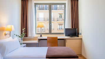 Habitación de hotel con cama, escritorio, silla, televisor y ventana con cortinas.