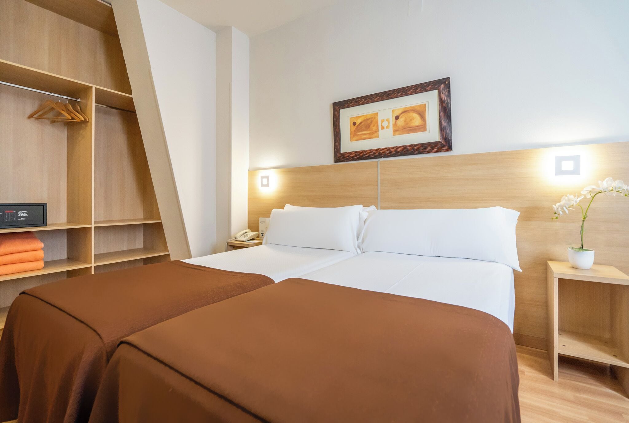 A hotel room with two beds, a wardrobe, a bedside table with a lamp, and a framed picture on the wall.