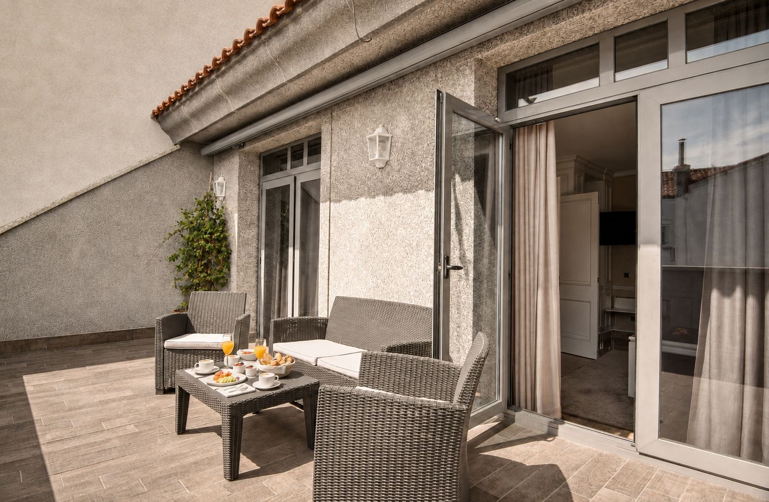 Une terrasse dotée de meubles en osier, une table mise pour le petit-déjeuner et une porte-fenêtre coulissante donnant sur un espace intérieur.