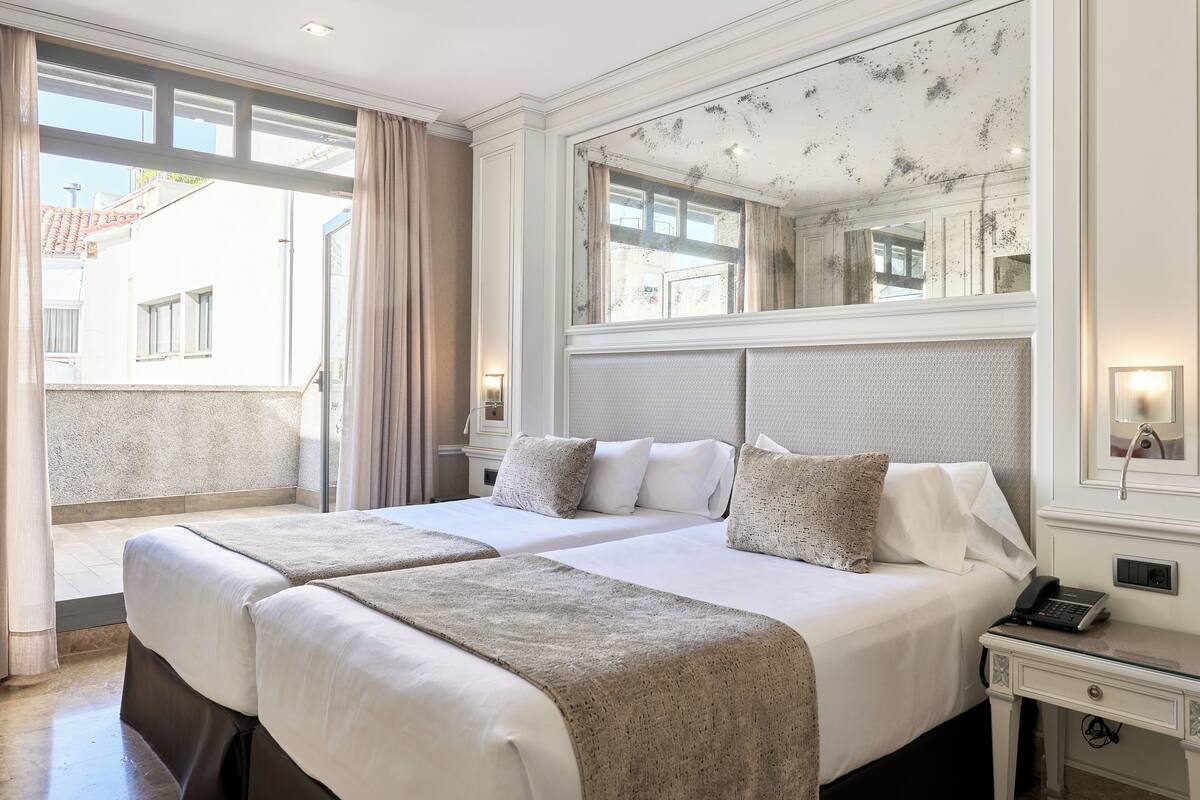 Ein Hotelzimmer mit zwei Betten, einem großen Spiegel, einem Nachttisch mit Lampe und Blick nach außen.
