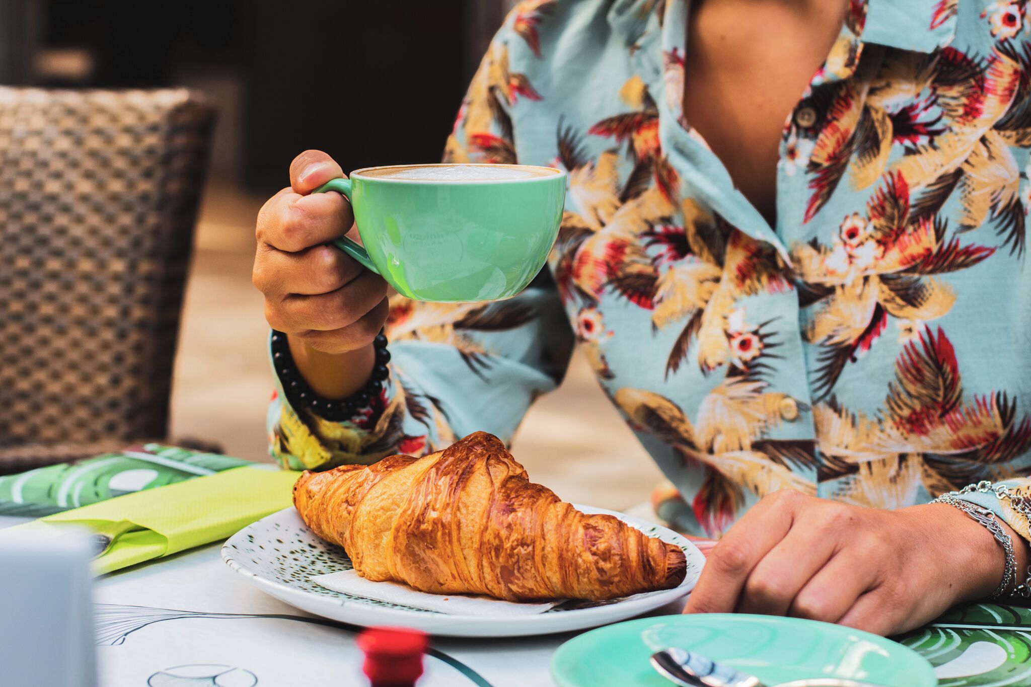 A person holding a green cup, sitting at a table with a croissant and a plate.