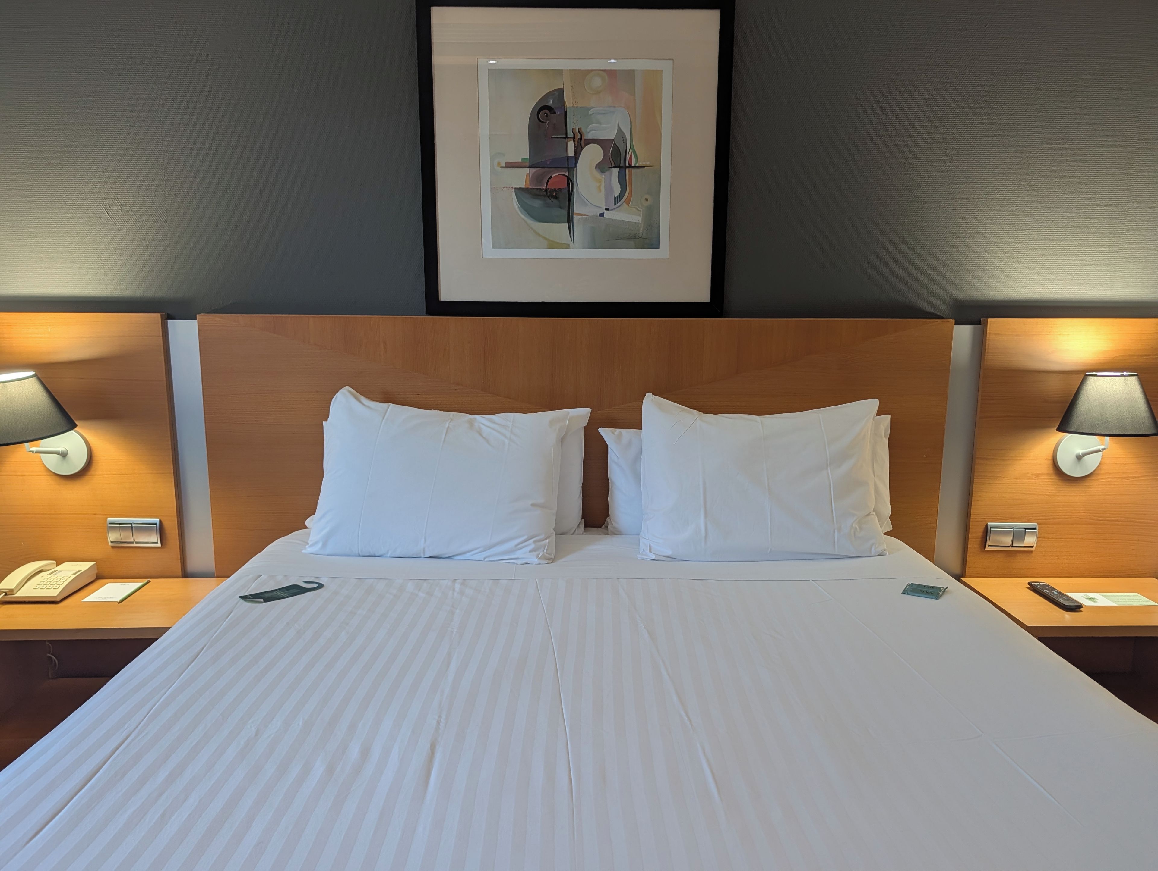 Habitación de hotel con una cama grande, dos lámparas de noche, una obra de arte enmarcada sobre la cama y un cabecero de madera.