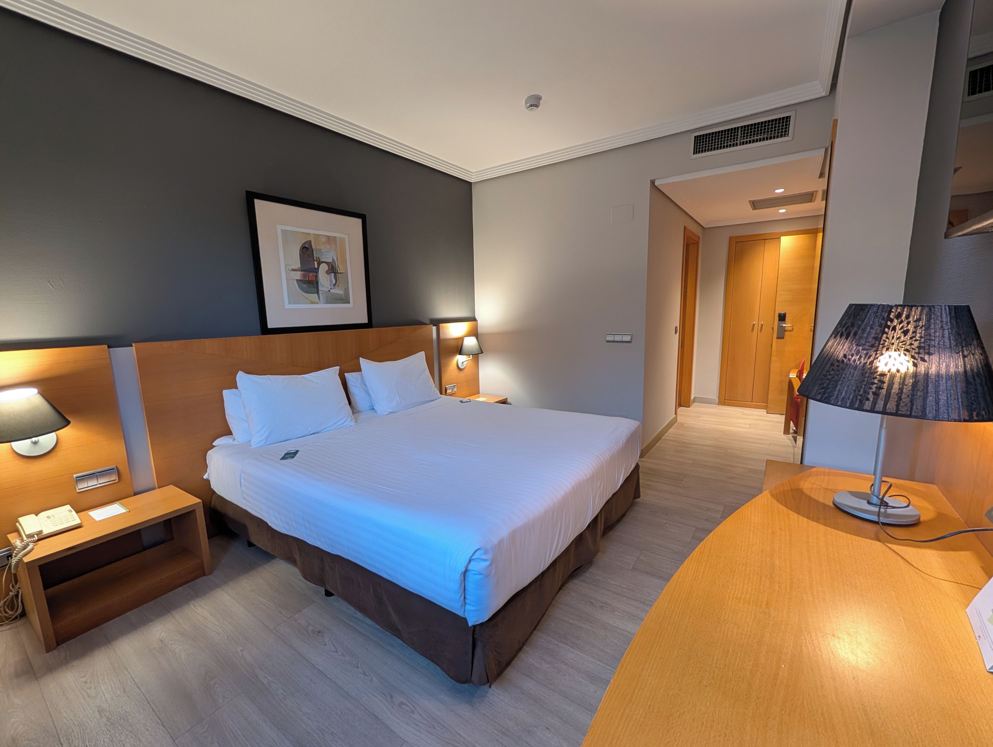 Ein Hotelzimmer mit einem großen Bett, Nachttischlampen, einem Holznachttisch, einem Schreibtisch mit Lampe und einem gerahmten Bild an der Wand.