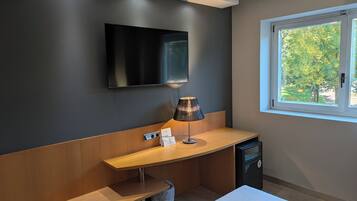 Une chambre d’hôtel moderne équipée d’un téléviseur à écran plat fixé au mur, d’une table de chevet avec une lampe, d’une poubelle et d’une fenêtre donnant sur des arbres.