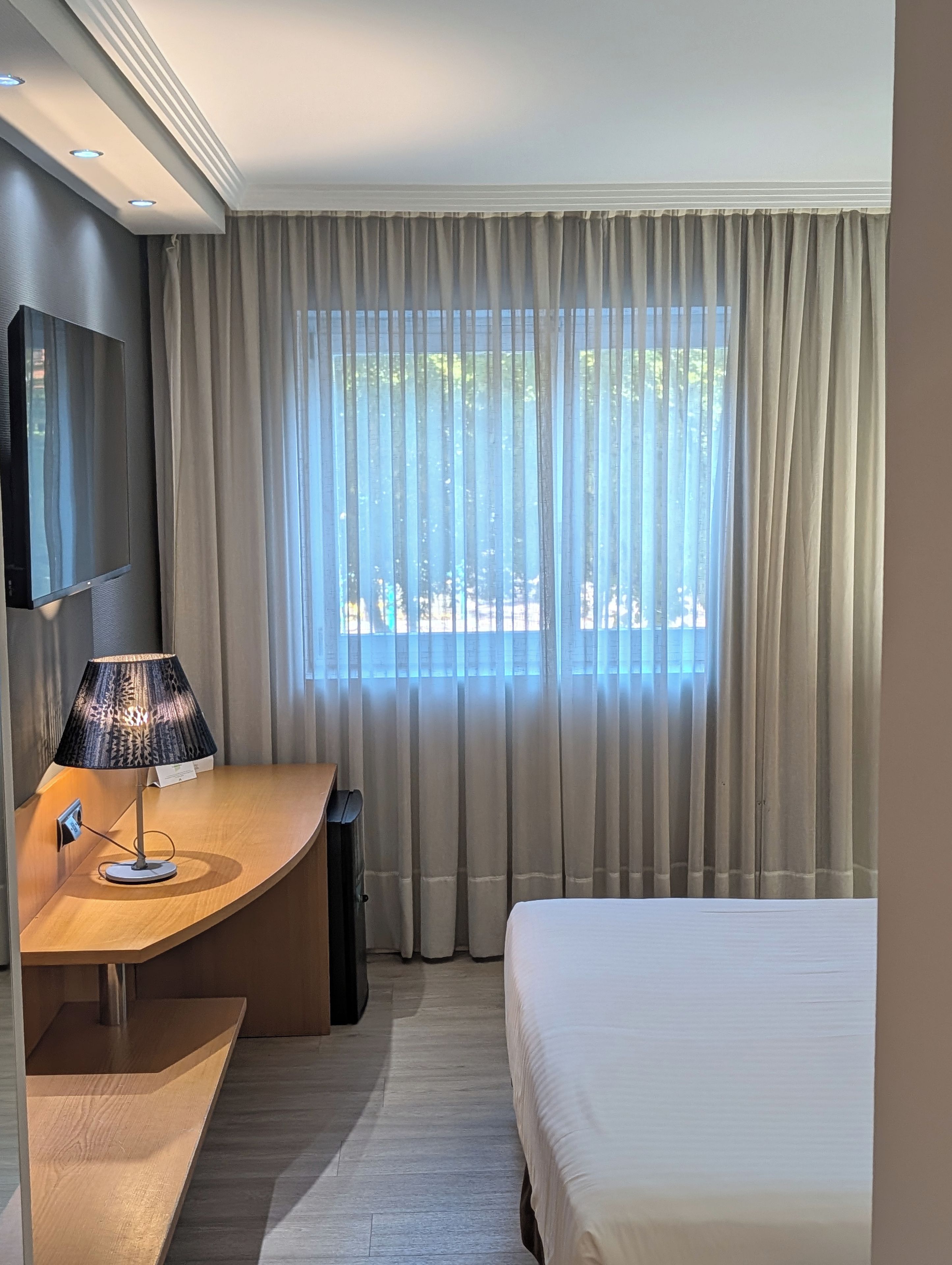 A hotel room with a bed, a desk with a lamp, a TV, and a window with curtains.