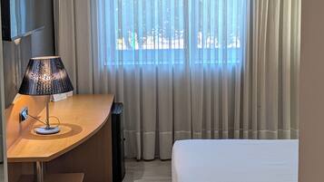 A hotel room with a bed, a desk with a lamp, a TV, and a window with curtains.