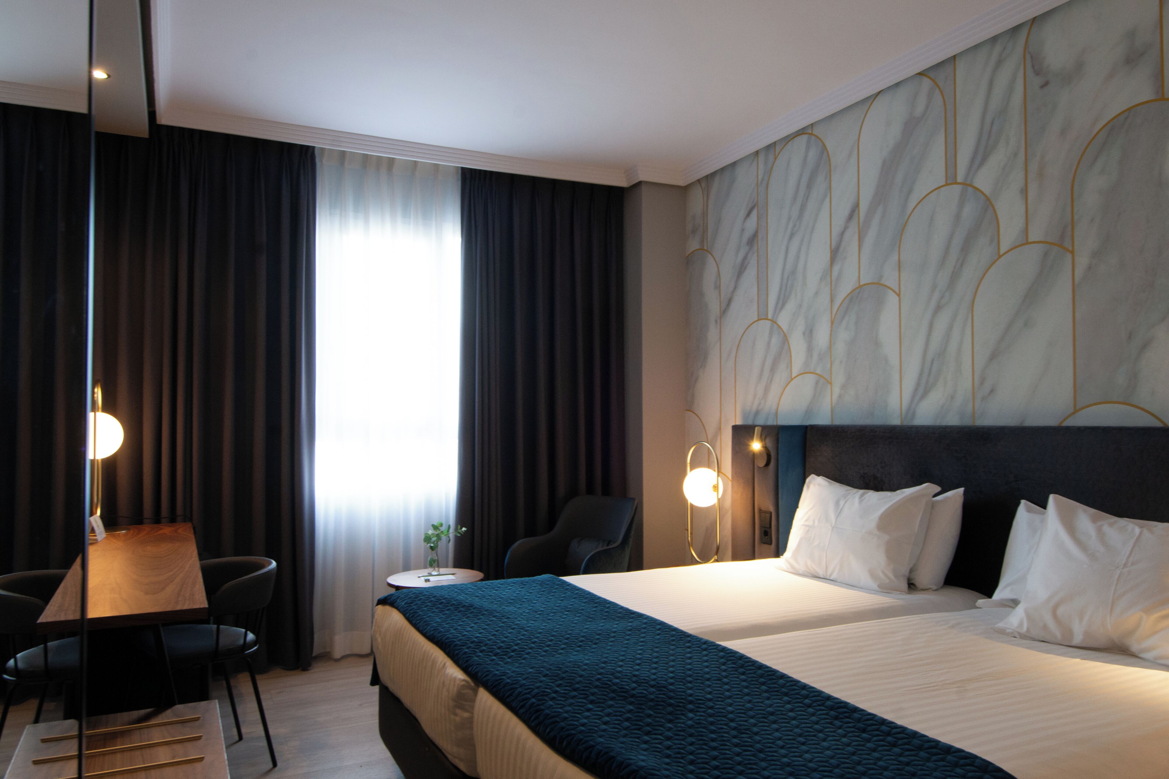 Habitación de hotel con cama, escritorio, silla y un amplio ventanal con cortinas.