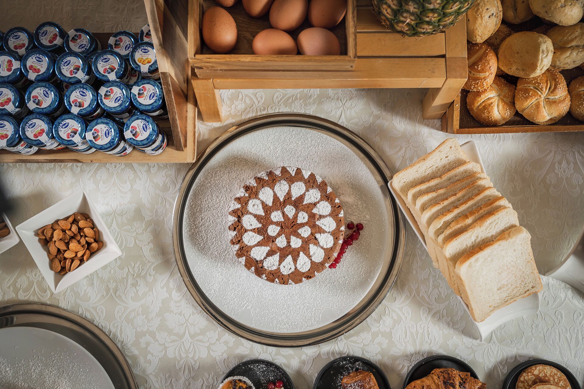 A top-down view of a kitchen counter with various food items, including a pie with a decorative crust, sliced bread, eggs, and jars of jam.