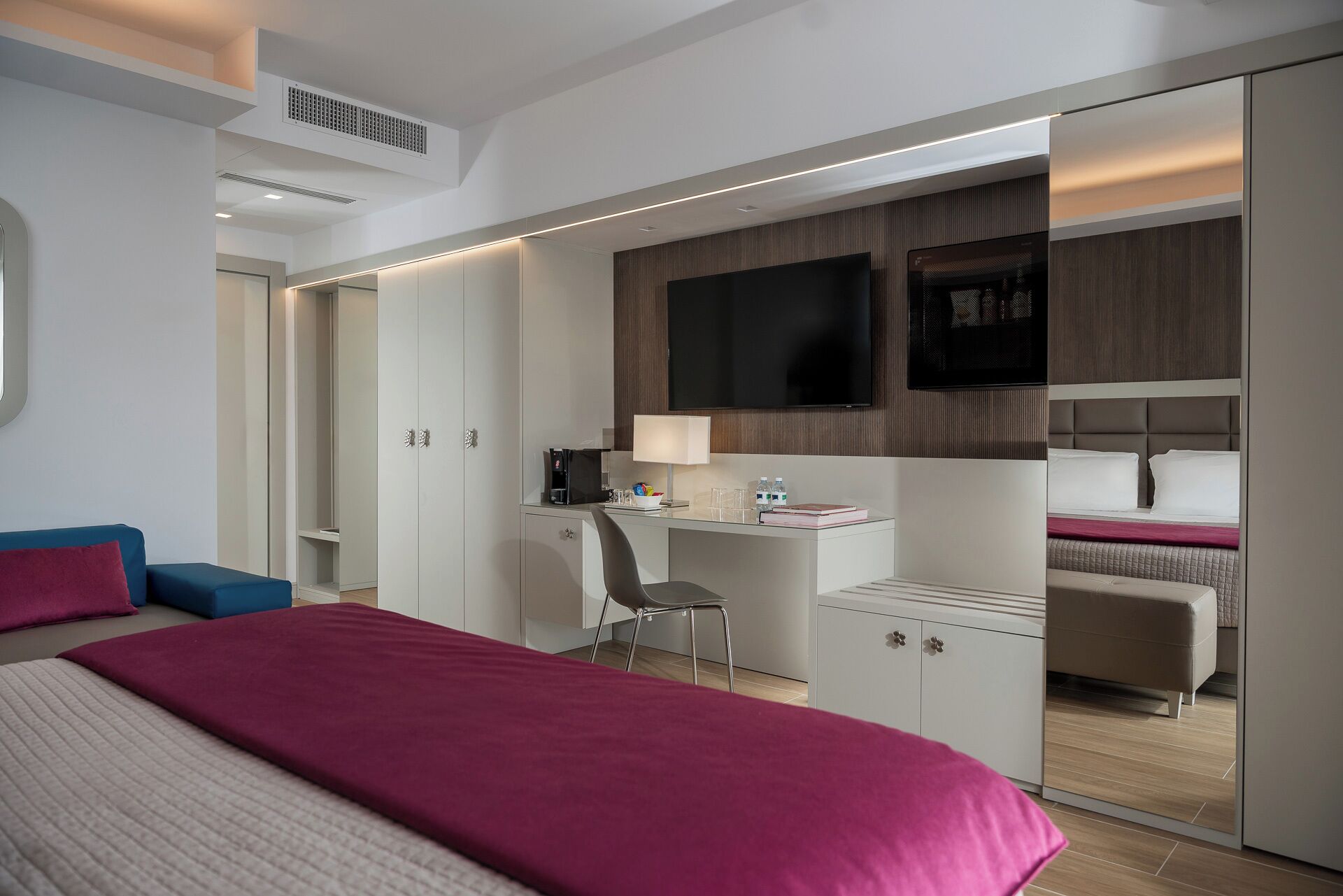 A modern hotel room with a large bed, a desk with a chair, a television, and a mirrored wardrobe.