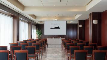 A conference room with rows of wooden chairs, a large screen displaying 'GRAND HOTEL SAN MARINO GROUP', and a potted plant.