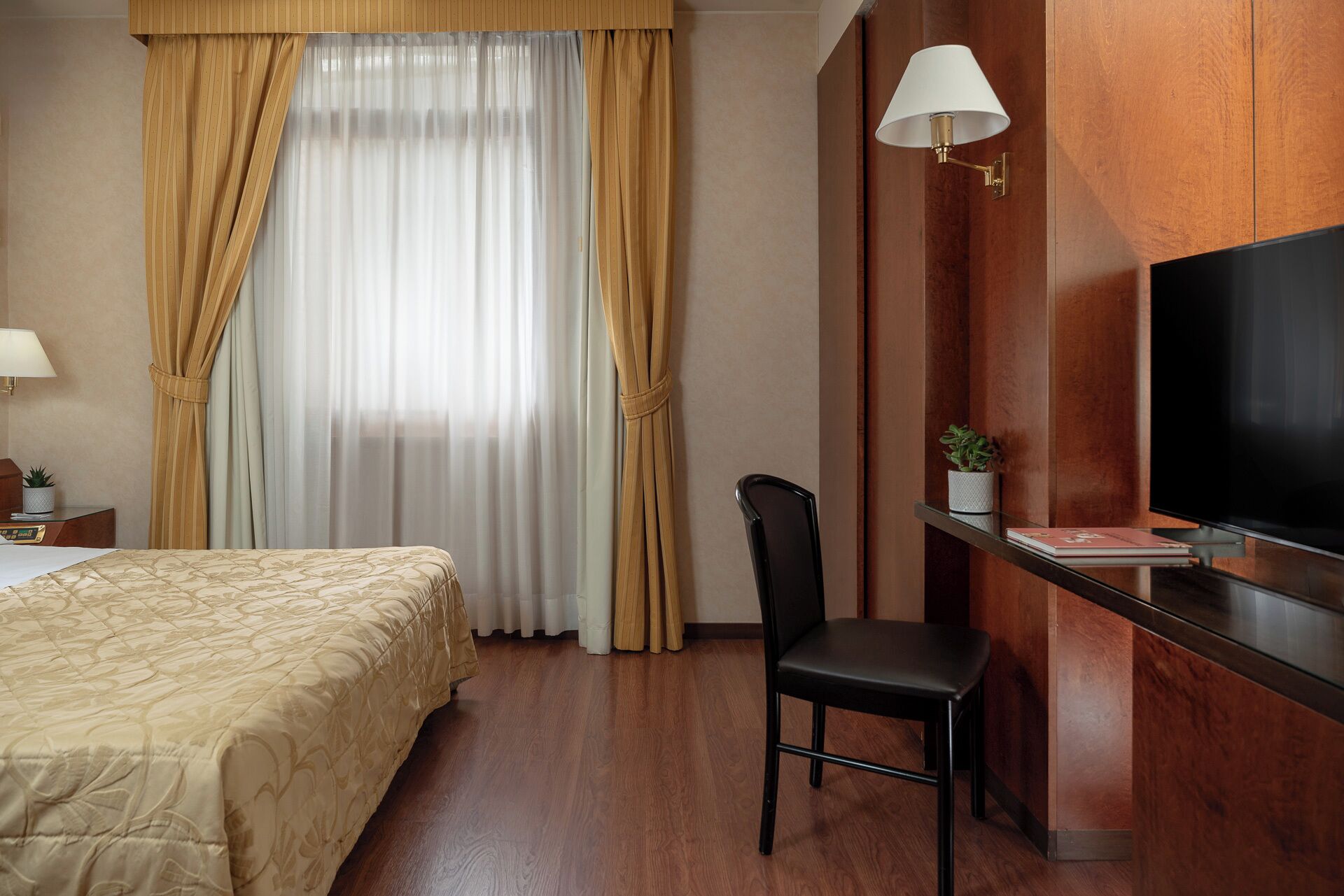 A hotel room with a bed, a desk with a chair, a TV, and a window with curtains.
