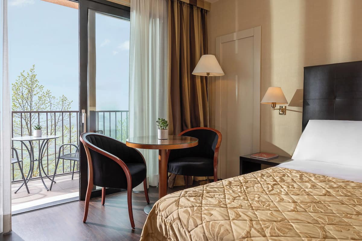 A hotel room with a bed, a small table with two chairs, a nightstand, and a view of trees through a balcony.