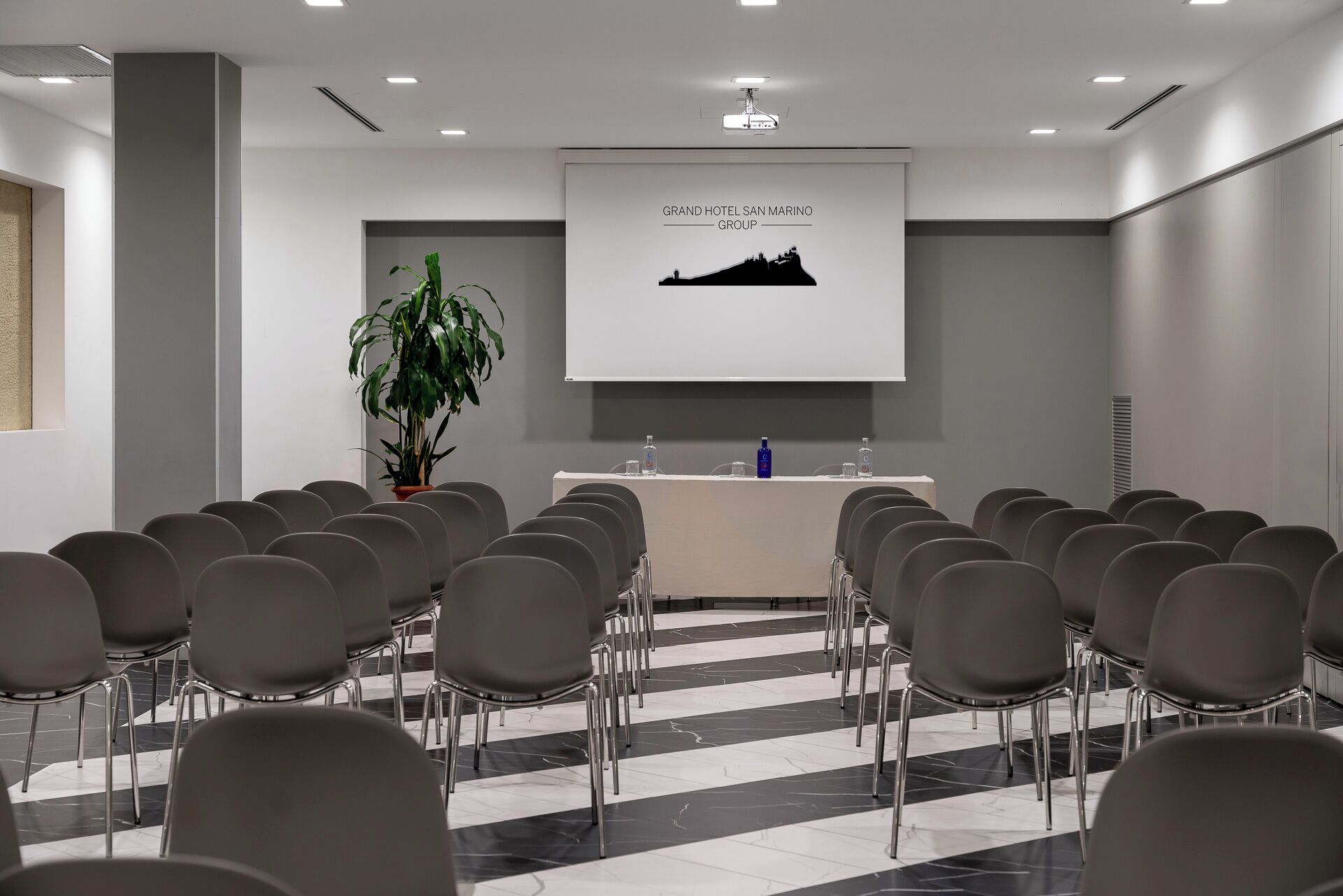 Una sala de conferències amb una taula llarga, cadires, una pantalla de projector i una planta en test.