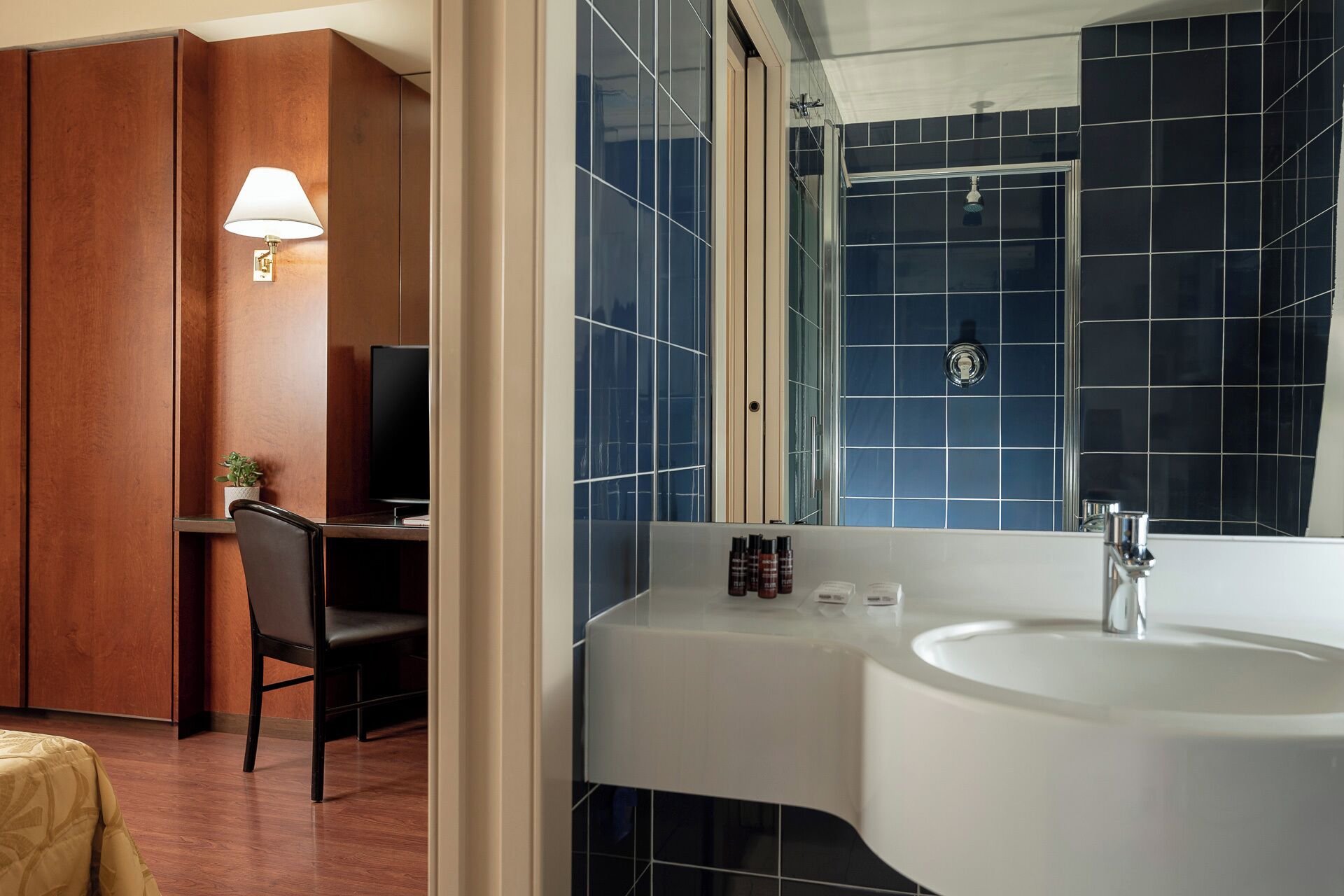 Un baño de hotel con un lavamanos grande, un espejo y una ducha. En la habitación contigua hay una silla y un televisor.
