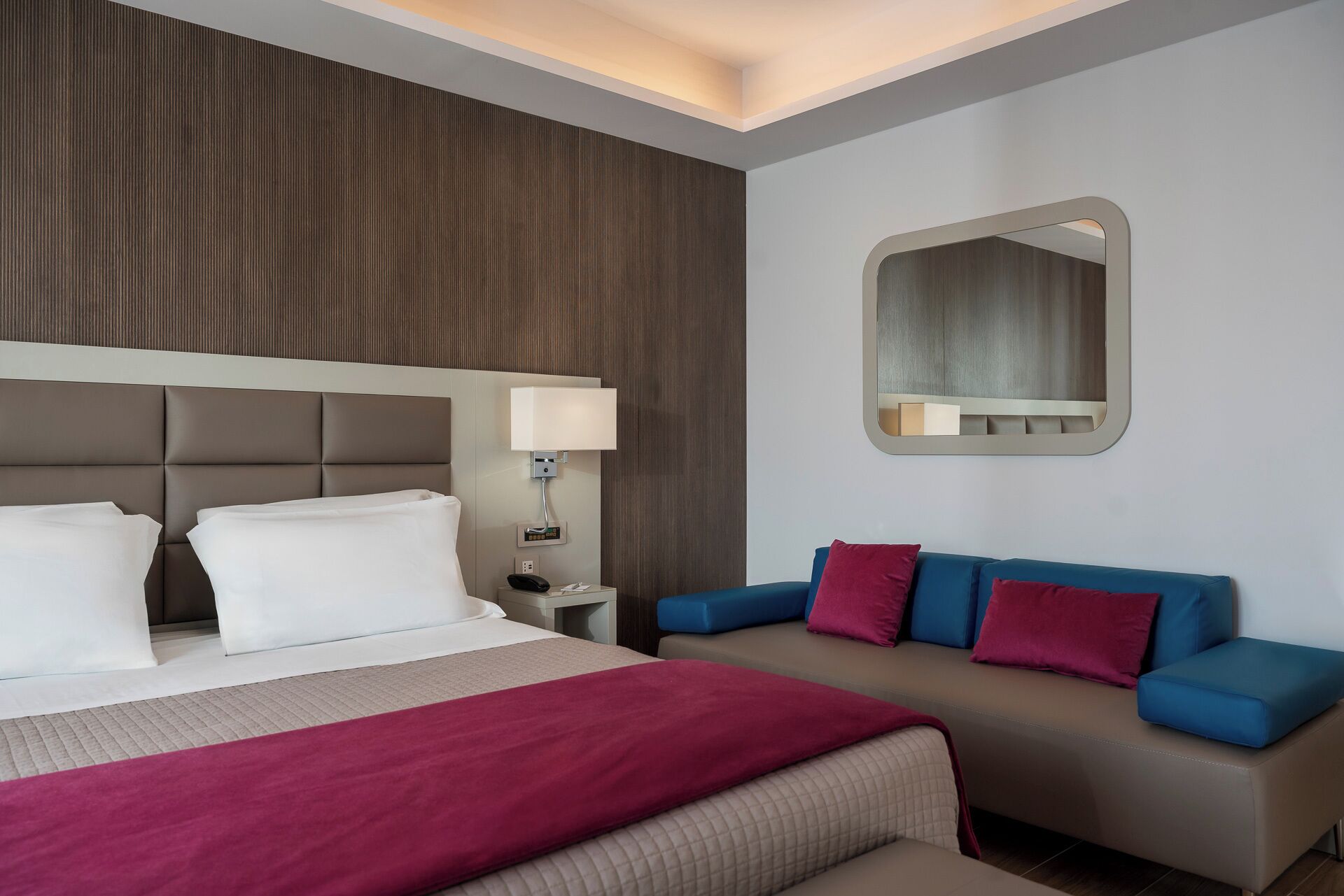 A hotel room with a bed, a nightstand, a lamp, a sofa, and a mirror.