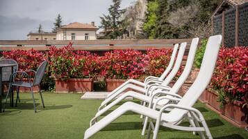 A rooftop terrace with white lounge chairs and a table, surrounded by red flowering plants.