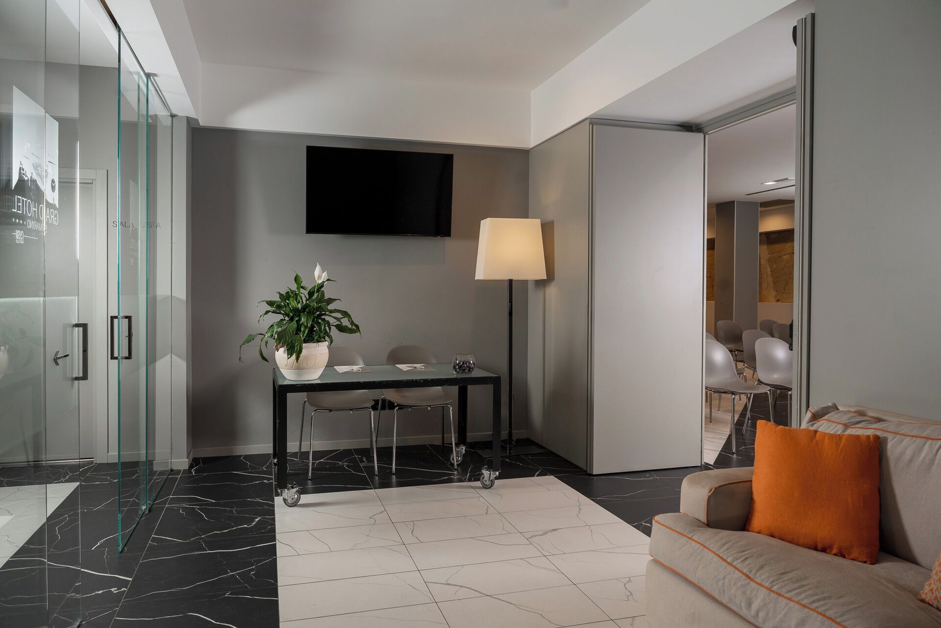Una habitación de hotel moderna con ducha de vidrio, un televisor de pantalla plana montado en la pared, un escritorio con una planta, una lámpara de pie y un sofá con un cojín naranja.