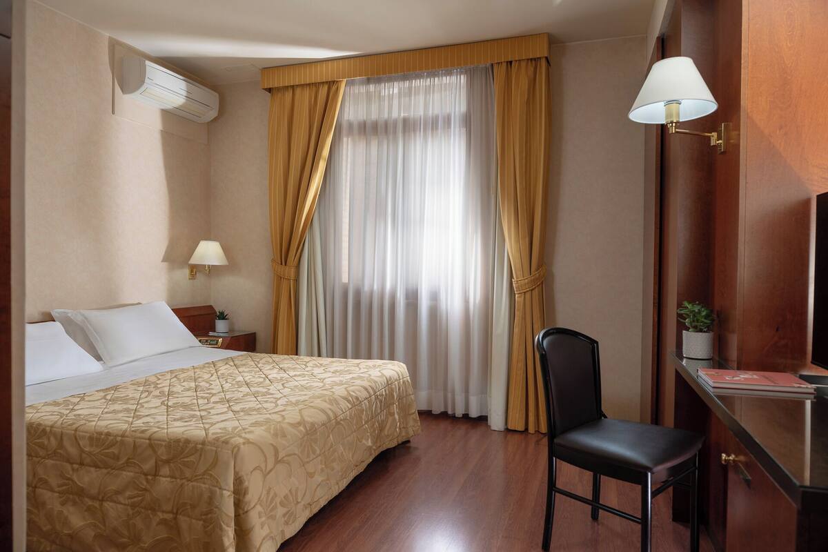 A hotel room with a bed, a desk with a lamp, a chair, a TV, and a window with curtains.