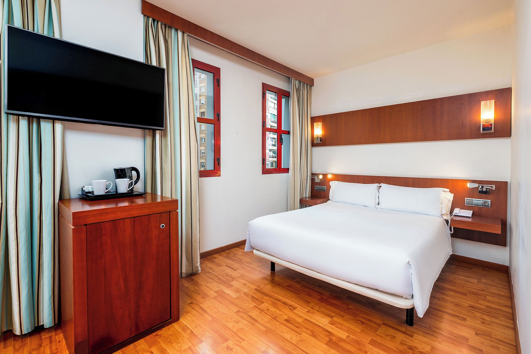 A hotel room with a bed, a TV, a bedside table with a lamp, a wooden desk, and curtains.