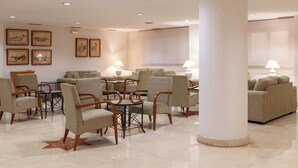 Un vestíbulo espacioso con varias zonas de estar, que incluyen sillones y sofás, distribuidos alrededor de una mesa central. Hay obras de arte enmarcadas en las paredes y un pilar grande en el centro.