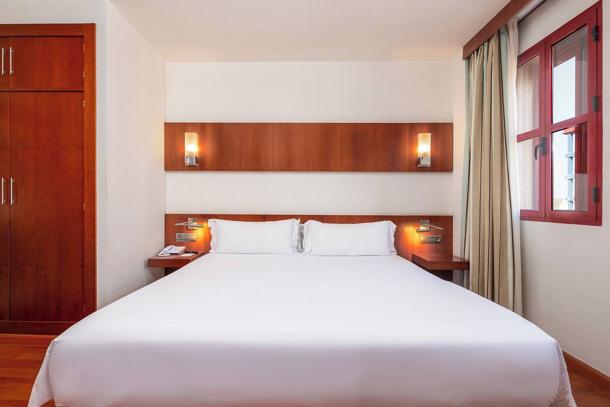 A hotel room with a large bed, wooden headboard, bedside lamps, and a wardrobe.