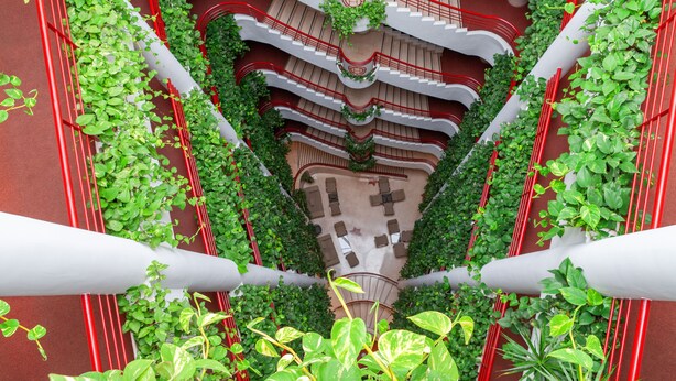 Un atrio de varios niveles con barandillas rojas, plantas verdes y un espacio central abierto con asientos.