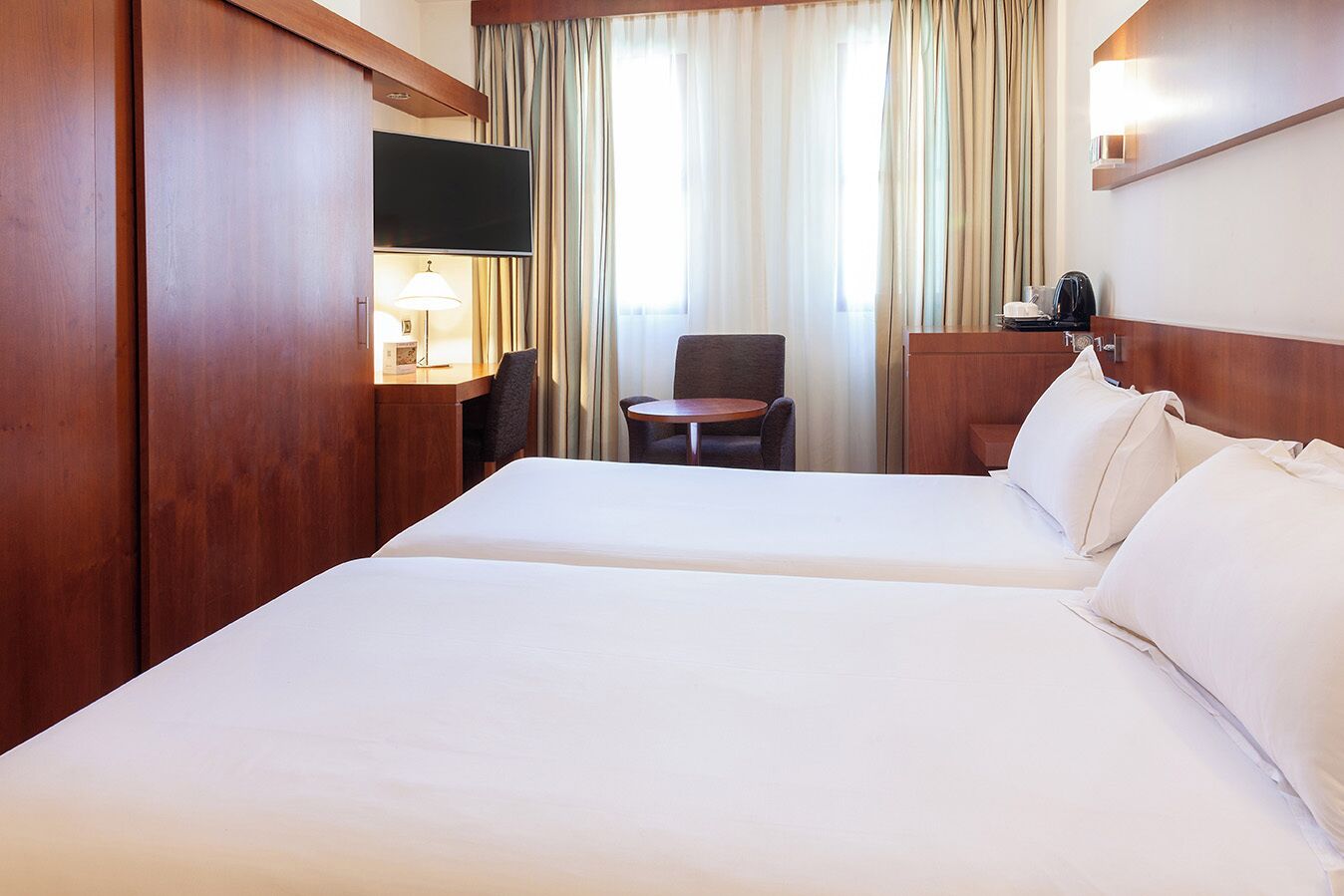 A hotel room with two beds, a desk with a lamp, a chair, and a TV.