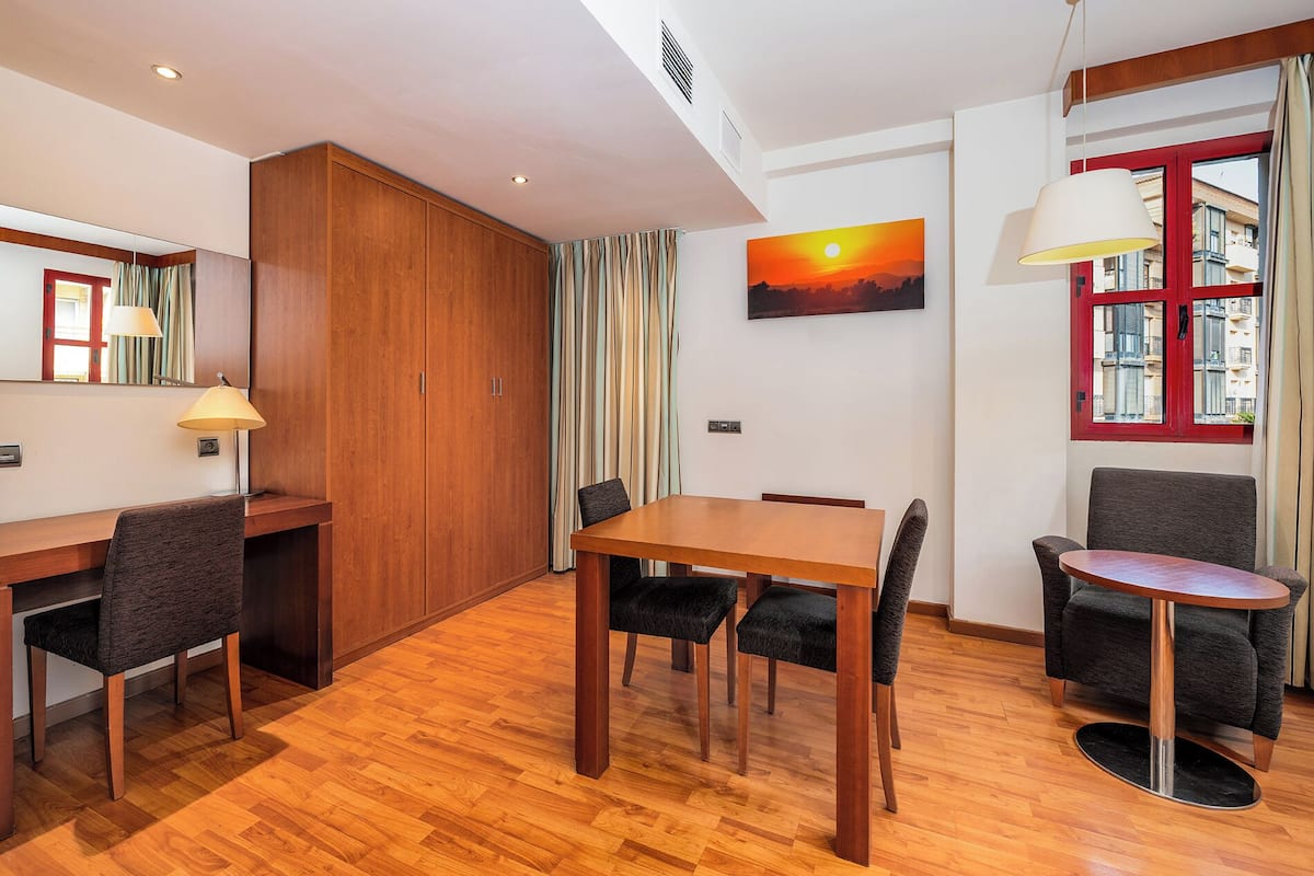 A modern hotel room with a wooden floor, a dining table with four chairs, a small round table with a lamp, a large window with a view of buildings, and a painting of a sunset on the wall.
