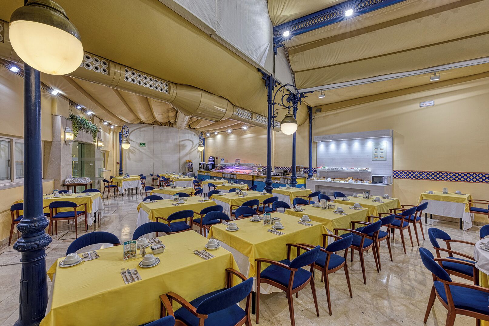 Een ruime eetruimte met gele tafels en blauwe stoelen, een buffet en een betegelde vloer.
