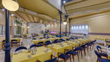 Un comedor amplio con mesas amarillas y sillas azules, un mostrador de buffet y piso de baldosas.