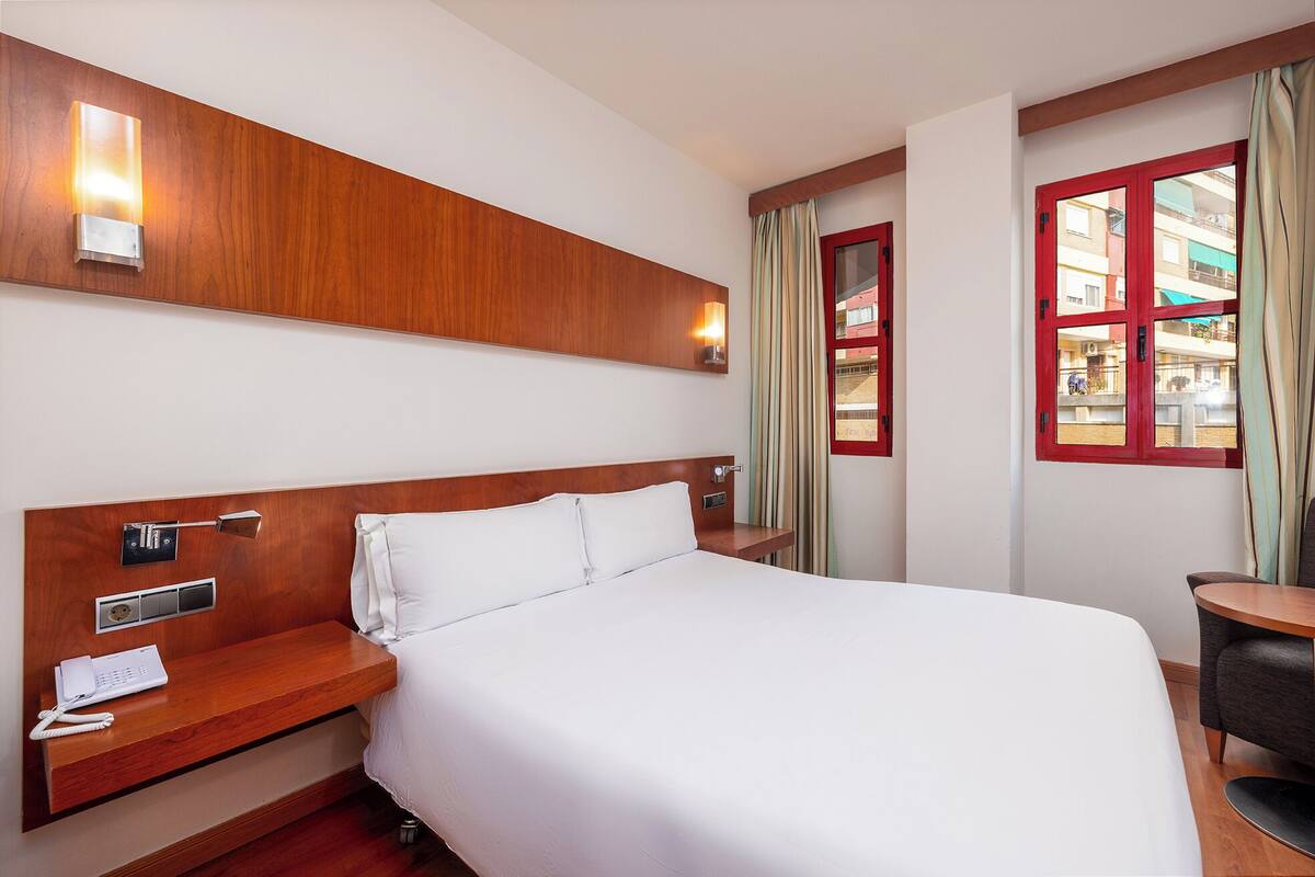 A hotel room with a wooden headboard, a bedside table with a telephone, a window with curtains, and a view of buildings outside.