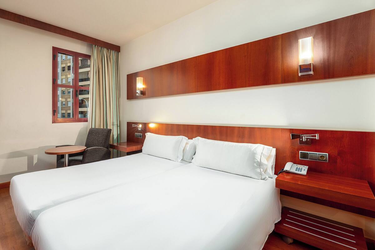 A hotel room with a large bed, a bedside table with a lamp, a chair, and a window with curtains.