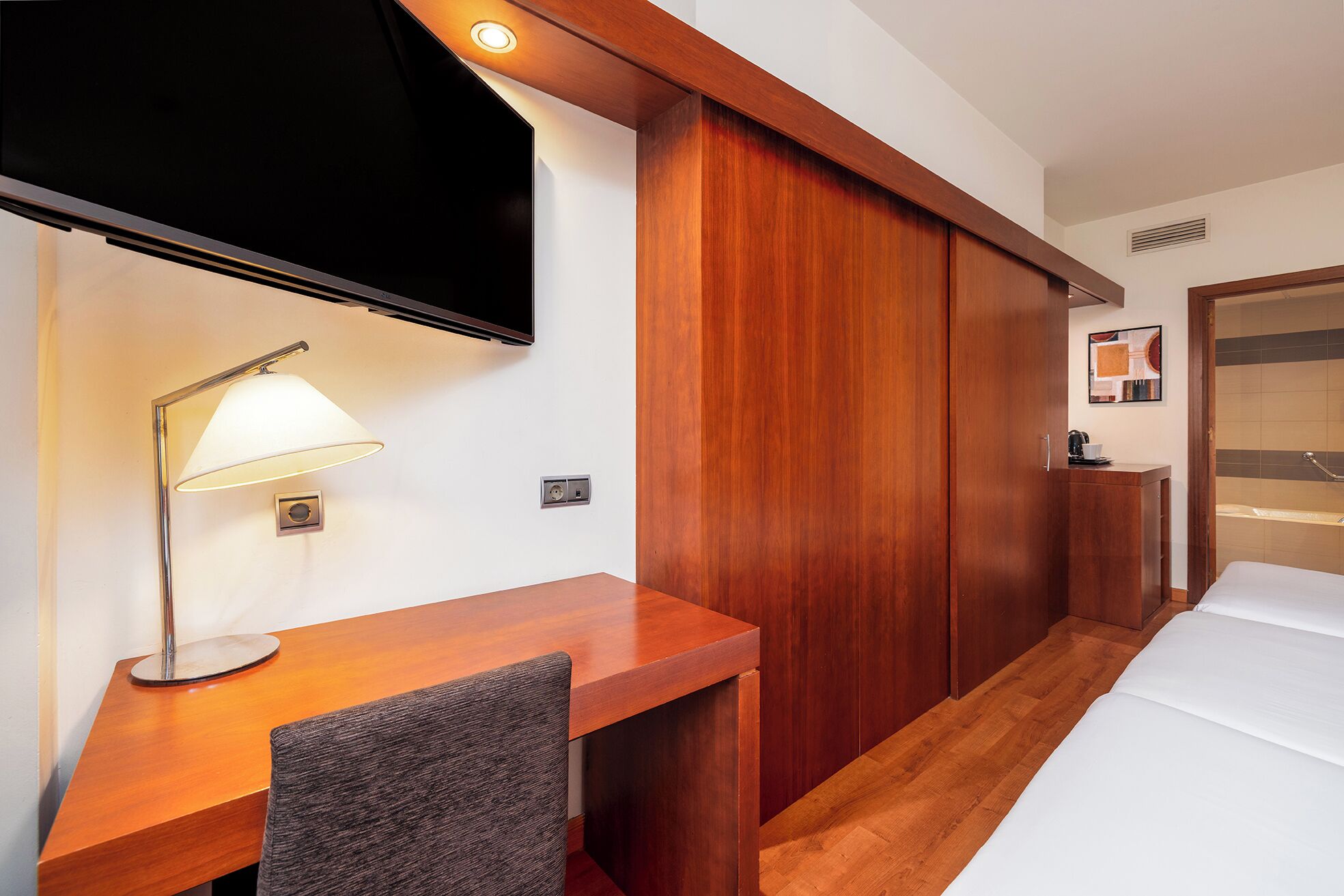 A hotel room with a wooden desk, a TV mounted above it, a bedside table with a lamp, a wardrobe, and a bed.