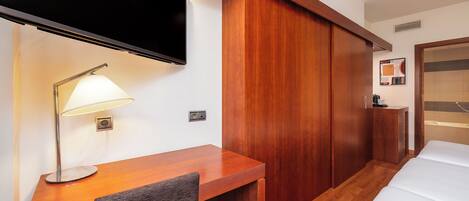 A hotel room with a wooden desk, a TV mounted above it, a bedside table with a lamp, a wardrobe, and a bed.