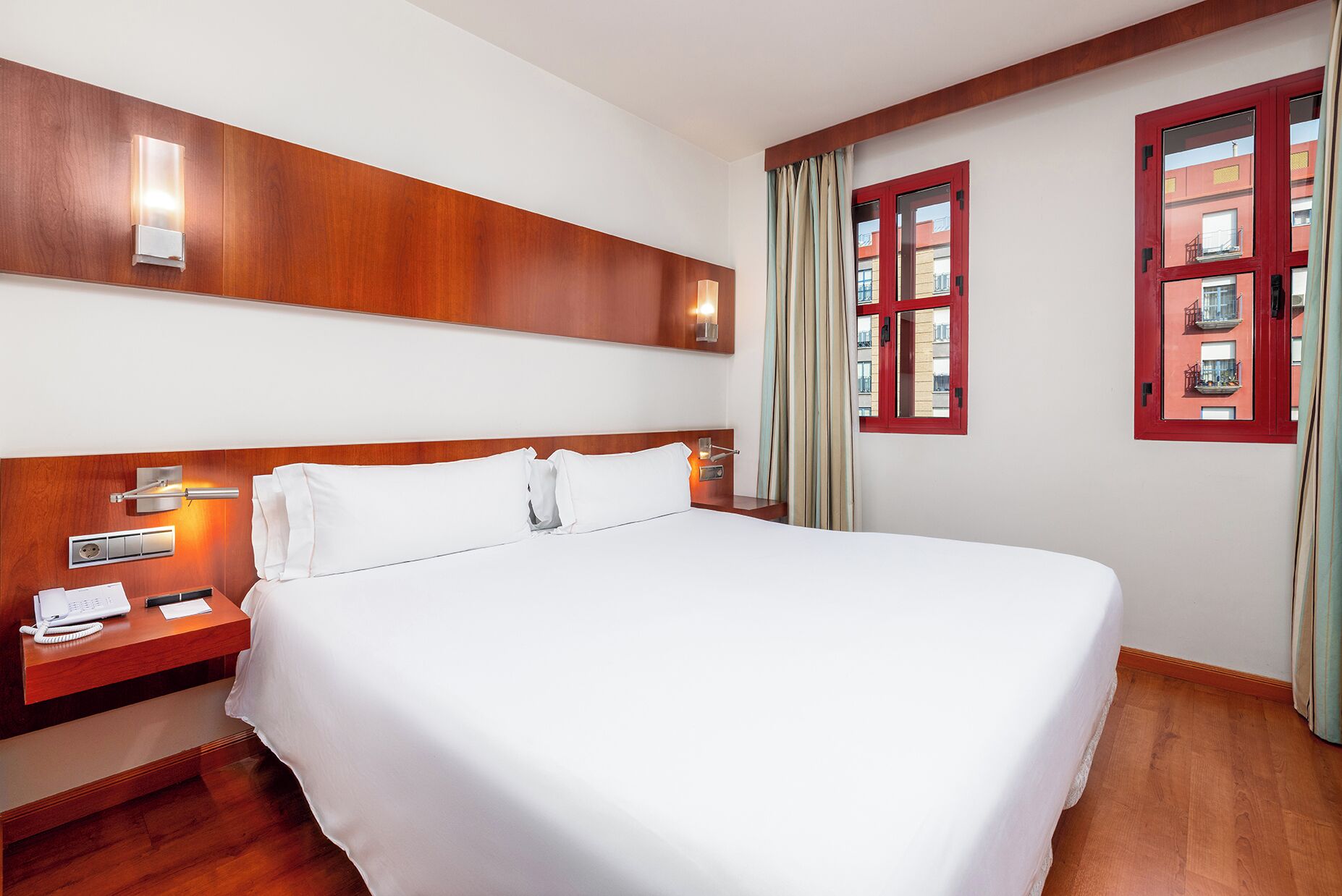 Habitación de hotel con una cama grande, cabecera de madera, mesitas de noche con lámparas, un teléfono y una ventana con cortinas.