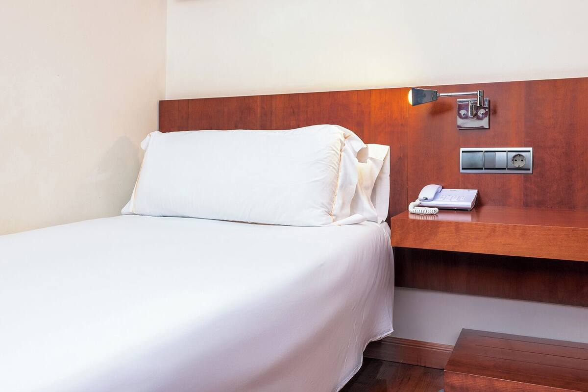 A hotel room with a neatly made bed, a bedside table with a lamp, a phone, and a power outlet.