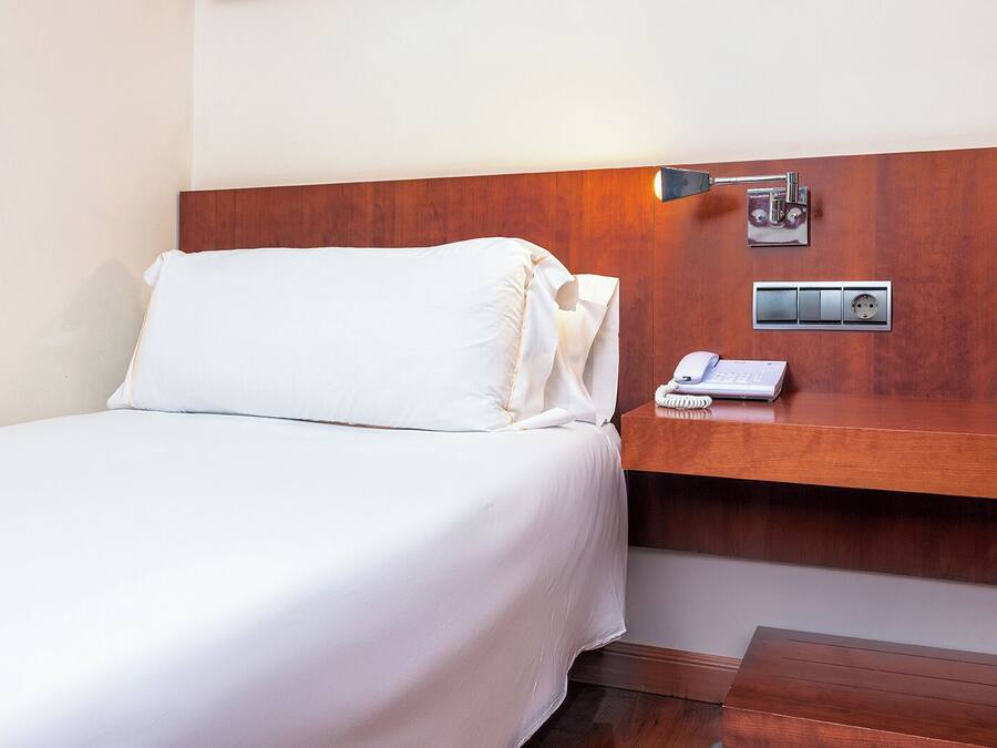 Une chambre d’hôtel comprenant un lit, une table de chevet avec un téléphone et une prise électrique, ainsi qu’un luminaire fixé au mur.