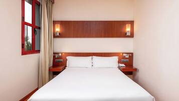 A hotel room with a large bed, wooden headboard, two bedside tables with lamps, and a window with curtains.