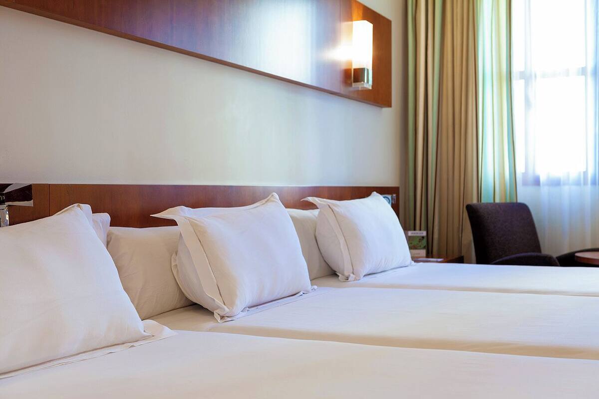 A hotel room with two beds, a wooden headboard, a wall-mounted light, and a view of the outside through curtains.