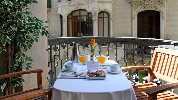 An outdoor dining table set with breakfast items, a flower, and a view of ornate building architecture. - Hotel Nouvel (Barcelona)